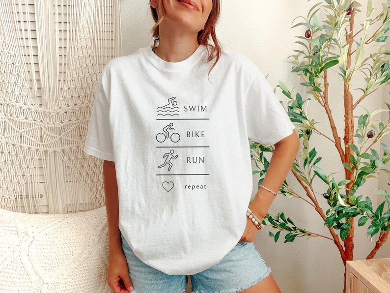May include: White t-shirt with a black graphic design. The design features the words "swim, bike, run, repeat" with corresponding illustrations of a swimmer, cyclist, runner, and a heart. The shirt is worn with blue denim shorts.