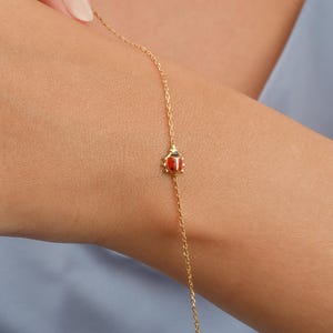 Peut inclure: Un bracelet en or délicat avec un petit charme de coccinelle avec des détails en émail rouge et noir. Le bracelet est une fine chaîne, et le charme de coccinelle est centré sur le poignet. Le fond est bleu clair.
