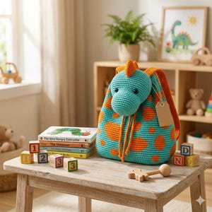 Könnte beinhalten: Ein türkisfarbener und orangefarbener Häkelrucksack in Dinosaurierform mit rundem Kopf, kleinem Kamm und orangefarbenen Punkten. Der Rucksack hat orangefarbene Träger, einen Kordelzug und ein leeres beiges Papieretikett an der Seite.