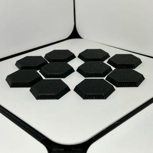 May include: A collection of ten black, hexagonal tiles arranged on a white surface. Each tile has a textured surface and a beveled edge. The tiles are evenly spaced and appear to be made of a solid material.