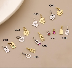 May include: A collection of gold and silver-toned charms, including initial letters, a heart, a puzzle piece, and a flower. Some charms feature gemstones. The charms are designed for jewelry making.