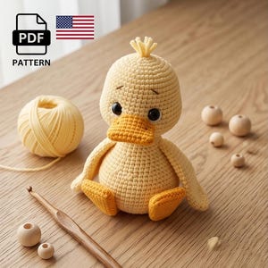 May include: A crocheted yellow duckling with an orange beak and feet sits on a wooden surface. A ball of yellow yarn, a crochet hook, and wooden beads are also present. The image includes a PDF pattern icon and the American flag.