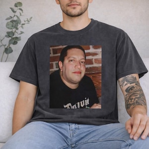 May include: Dark gray t-shirt featuring a framed photograph of a person. The photograph is of a man in a black shirt. The t-shirt has a vintage, washed look.