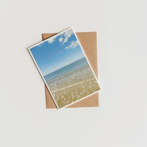 May include: A photograph of a beach scene with blue sky and ocean waves, resting on a brown envelope. The image is angled, showing the horizon line and the sandy shore. The background is a plain white surface.