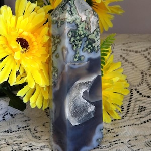 May include: A polished moss agate crystal tower featuring translucent grey and white bands with distinct green moss-like inclusions and a small, textured crystalline geode cavity visible on the front face of the stone pillar.
