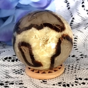 May include: A spherical gemstone with a cream and brown pattern. The orb is displayed on a small, light brown wooden stand. The background features a white lace cloth and a blue flower.