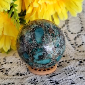 May include: A polished spherical stone featuring a marbled pattern of deep teal, turquoise, and dark brown veins. The sphere rests on a small, circular, light-coloured wooden stand with a cutout geometric pattern, showcasing a smooth, glossy surface.