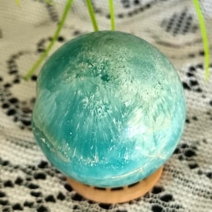 May include: A polished spherical stone featuring a vibrant teal and turquoise colour palette with intricate white crystalline patterns radiating from the centre. The sphere rests securely on a small, circular, light-coloured wooden display stand.