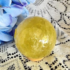 May include: A polished, translucent yellow sphere made of natural crystal, resting on a small wooden base. The sphere displays internal fractures and inclusions, reflecting light across its smooth, rounded surface with a bright, sunny hue.