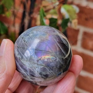 May include: A polished spherical labradorite gemstone featuring a dark grey base with prominent internal fractures and a vibrant iridescent flash of purple, blue, and gold light reflecting across its smooth, rounded surface.