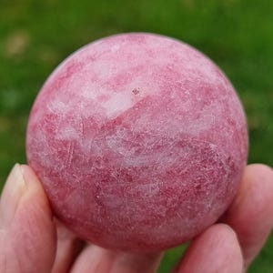 May include: A polished spherical gemstone featuring a vibrant pink colour with intricate white veining and natural crystalline textures throughout the surface. The stone displays a smooth, rounded shape with subtle variations in shade and depth.