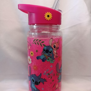 May include: A clear plastic water bottle with a pink glitter liquid interior, featuring a pink lid with a clear straw and a yellow flower accent. The exterior is decorated with a repeating pattern of blue and pink alien characters and flowers.