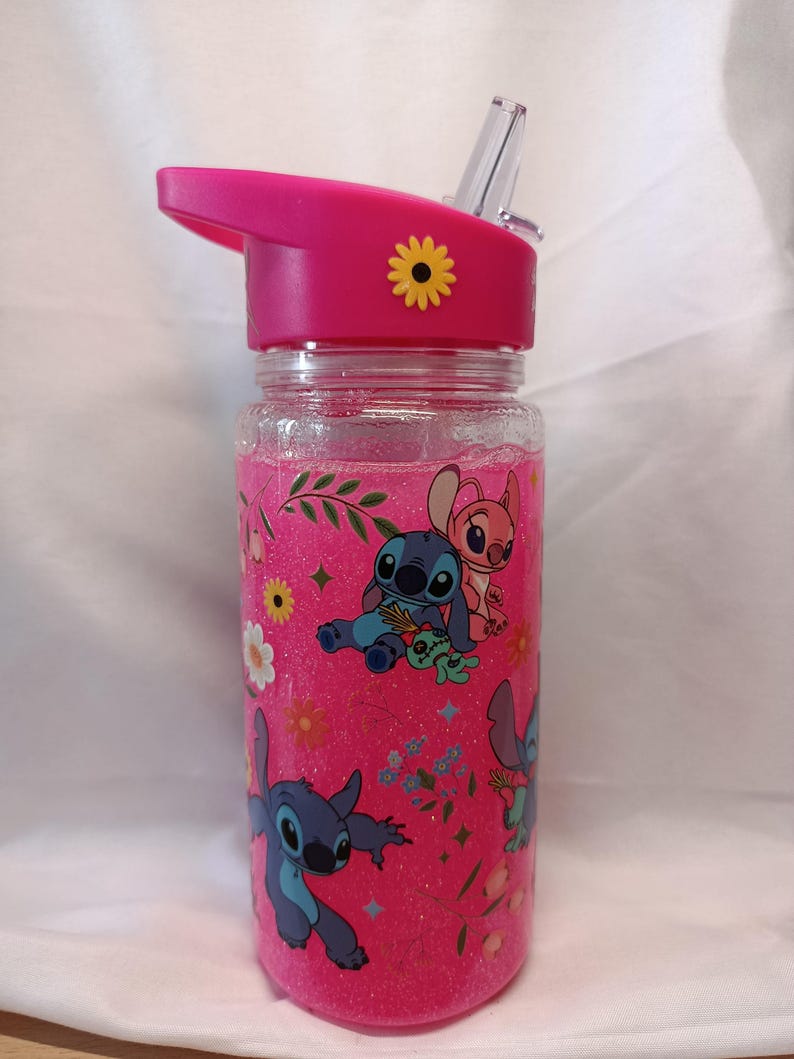 May include: A clear plastic water bottle with a pink glitter liquid interior, featuring a pink lid with a clear straw and a yellow flower accent. The exterior is decorated with a repeating pattern of blue and pink alien characters and flowers.
