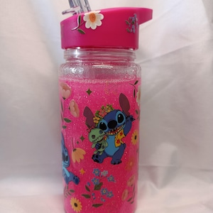 May include: A clear plastic water bottle filled with pink glitter liquid, featuring a bright pink lid with a straw. The bottle is decorated with floral patterns and a graphic of the blue character Stitch holding a small green doll.
