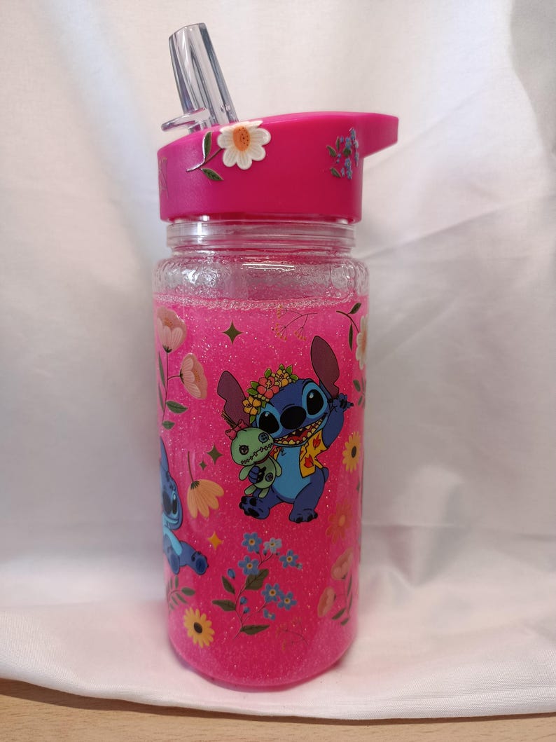 May include: A clear plastic water bottle filled with pink glitter liquid, featuring a bright pink lid with a straw. The bottle is decorated with floral patterns and a graphic of the blue character Stitch holding a small green doll.