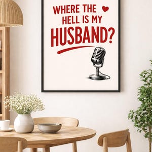 May include: A framed print with the text "WHERE THE HELL IS MY HUSBAND?" in red, with a heart symbol and a vintage microphone illustration. The print is in a black frame, displayed in a room with a wooden table and chairs.