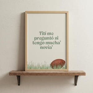 Op de afbeelding: Ingelijste print met de tekst "Titi me preguntó si tengo mucha' novia" in het groen. De print toont een voetbal en gras aan de onderkant. De lijst is lichtbruin en wordt op een houten plank gepresenteerd.