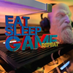 Könnte beinhalten: Ein 3D-gedrucktes Schild mit den Worten "EAT SLEEP GAME REPEAT" in Blau, Rot und Gelb. Das Schild befindet sich auf einer schwarzen Oberfläche, mit einer Büste eines bärtigen Mannes im Hintergrund. Es ist ein dekorativer Artikel zum Thema Gaming.