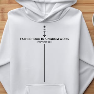 May include: A white hooded sweatshirt featuring a black graphic design on the front. The text reads FATHERHOOD IS KINGDOM WORK PROVERBS 22:6, accompanied by a vertical line and small cross symbols arranged to form a cross shape on the chest.