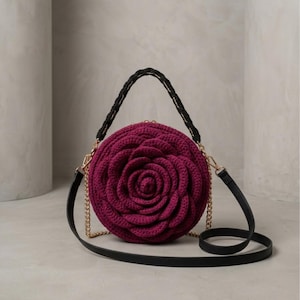 May include: A round, crocheted bag in a deep magenta color, shaped like a rose. It has a black braided handle and a black shoulder strap with gold chain accents. The bag is on a neutral background.