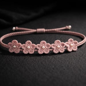 Puede incluir: Pulsera de ganchillo rosa con cinco aplicaciones de flores, cada una con una perla en el centro. La pulsera es ajustable con un nudo corredizo y tiene detalles de perlas en los extremos. Un accesorio encantador y floral.