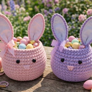 May include: Two handmade crochet Easter bunny baskets, one pink and one purple, each filled with colorful Easter eggs. The baskets have bunny ears and bows. A ball of pink yarn and a crochet hook are in the foreground.
