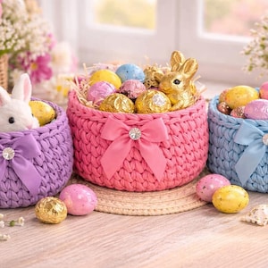 Könnte beinhalten: Drei farbenfrohe, gehäkelte Osterkörbe gefüllt mit dekorierten Eiern und Schokoladenbonbons. Die Körbe sind lila, rosa und blau, jeder mit einer Schleife und einer dekorativen Verzierung. Ein Korb enthält eine Hasenfigur.