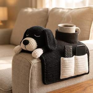 Könnte beinhalten: Ein schwarz-weiß gehäkelter Hunde-Sofa-Armlehnen-Organizer. Der Organizer hat zwei weiße Taschen, einen Halter für eine Tasse und einen Platz für eine Fernbedienung und ein Telefon. Eine Tasse Kaffee steht im Halter.
