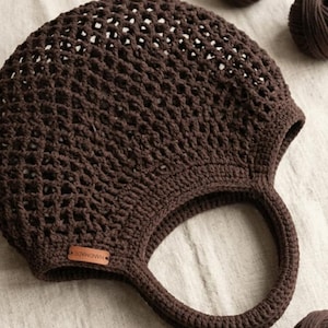 May include: A handmade, dark brown crochet bag with a round shape and two handles. The bag has an open weave design and a small leather tag that reads "HANDMADE". Three balls of brown yarn are next to the bag.