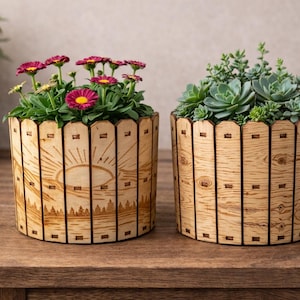 Puede incluir: Dos macetas de madera con plantas. Una contiene flores rosas y amarillas, la otra tiene suculentas verdes. Las macetas tienen un diseño de veta de madera clara con una escena de amanecer grabada con láser.