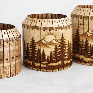 Könnte beinhalten: Drei dekorative Holzbehälter. Die Behälter sind zylindrisch mit hellbraunem Finish. Der größte Behälter zeigt eine Berg- und Waldszene mit einer Sonne. Der kleinste Behälter hat ein geometrisches Muster.