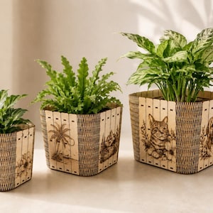Könnte beinhalten: Drei quadratische Pflanzgefäße aus Holz in verschiedenen Größen, jeweils mit lasergravierten Illustrationen von Tieren und Naturszenen. Die Gefäße haben eine helle Holzoberfläche mit dunklen, strukturierten vertikalen Paneelen.