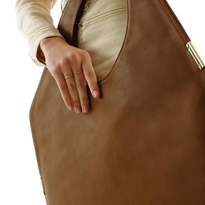 Op de afbeelding: Een bruine leren tote bag met een unieke driehoekige vorm. De tas heeft een grote opening en een goudkleurige metalen accent op de hoek. De tas wordt door een persoon vastgehouden.