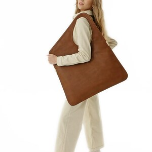 Op de afbeelding: Grote, vierkante bruine leren tote bag. De tas wordt vastgehouden door een persoon in een cr&egrave;mekleurige outfit. De tas heeft een minimalistisch design en een eenvoudige, strakke uitstraling, geschikt voor dagelijks gebruik.