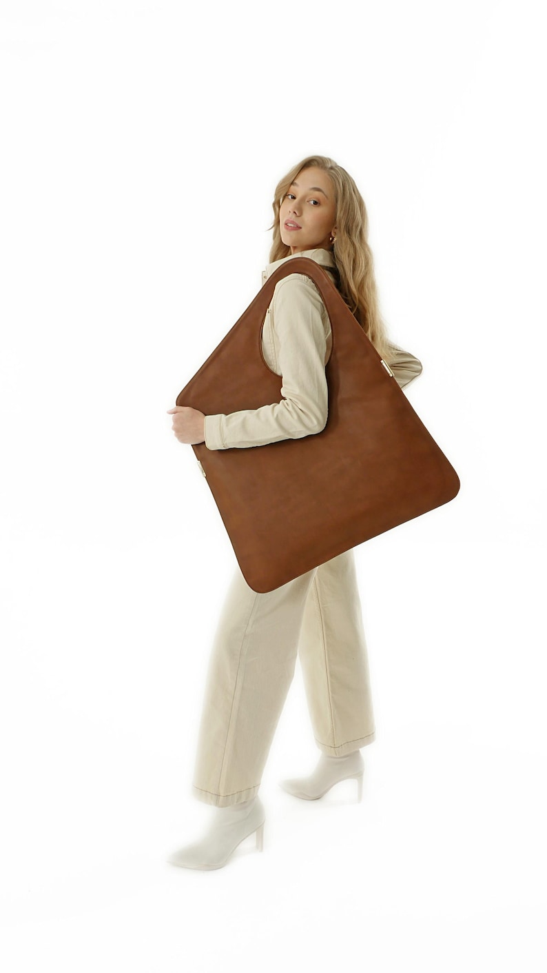 Op de afbeelding: Grote, vierkante bruine leren tote bag. De tas wordt vastgehouden door een persoon in een cr&egrave;mekleurige outfit. De tas heeft een minimalistisch design en een eenvoudige, strakke uitstraling, geschikt voor dagelijks gebruik.
