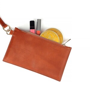 Op de afbeelding: Een bruine leren clutch met een riem, open om lipsticks en twee goudkleurige ronde items te laten zien. De clutch heeft een ritssluiting en een goudkleurige ringbevestiging voor de riem.