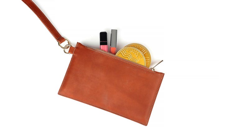 Op de afbeelding: Een bruine leren clutch met een riem, open om lipsticks en twee goudkleurige ronde items te laten zien. De clutch heeft een ritssluiting en een goudkleurige ringbevestiging voor de riem.