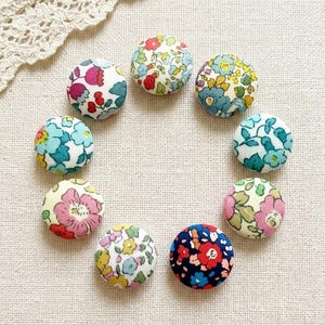 Può includere: Una collezione di nove bottoni rotondi rivestiti in tessuto con motivi floreali multicolori, tra cui rosso, blu, giallo e rosa. I bottoni sono disposti in cerchio su una superficie di tessuto color crema.