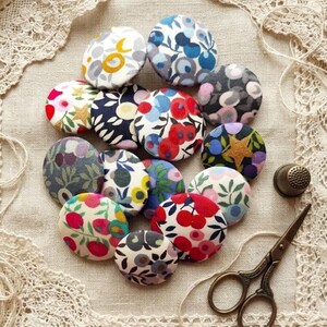Pode incluir: Uma coleção de botões coloridos revestidos de tecido com desenhos florais e estampados. Os botões estão dispostos sobre um tecido creme com acabamento em renda. Uma dedaleira e uma tesoura pequena também estão presentes.