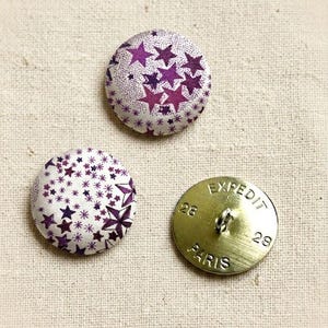 Pode incluir: Três botões redondos. Dois têm um padrão de estrelas roxas sobre fundo branco. O terceiro é de cor latão, com o texto "EXPEDIT PARIS" e os números 28 e 29.