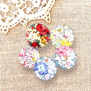 Pode incluir: Seis botões redondos cobertos de tecido com estampas florais em vermelho, rosa, amarelo e azul. Estão dispostos sobre um tecido bege texturizado. Um pedaço de renda branca é visível no canto superior esquerdo.