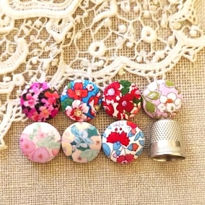 Puede incluir: Una colección de botones cubiertos de tela con estampados florales en varios colores, incluyendo rosa, rojo, azul y verde. Una dedal plateado se encuentra junto a los botones. Los botones están dispuestos sobre una tela beige.