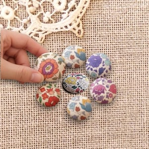 Pode incluir: Uma coleção de botões revestidos de tecido com padrões florais em várias cores, incluindo vermelho, roxo e azul. Os botões estão dispostos sobre uma superfície bege texturizada. Uma mão segura um dos botões.