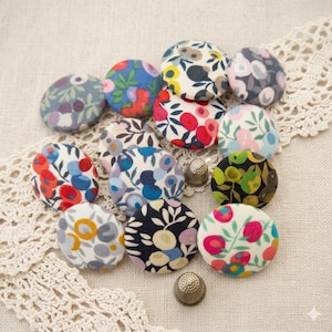 Puede incluir: Una colección de botones cubiertos de tela con estampados florales en varios colores, incluyendo azul, rojo, rosa y amarillo. Los botones están dispuestos sobre una superficie crema con ribete de encaje, y también se ven dos dedales.