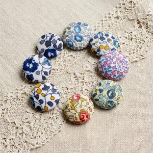 Può includere: Otto bottoni rivestiti in tessuto con motivi floreali, disposti a cerchio. I bottoni presentano disegni in blu, rosa, giallo e rosso. Ogni bottone misura circa 2,5 cm di diametro, su uno sfondo neutro.