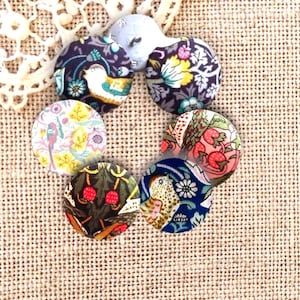 Pode incluir: Uma coleção de botões revestidos de tecido com padrões florais e de pássaros em várias cores, incluindo azul, rosa, amarelo e vermelho. Os botões estão dispostos em círculo sobre um fundo bege texturizado.