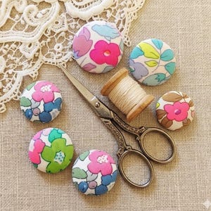 Pode incluir: Uma coleção de botões revestidos de tecido com padrões florais em rosa, azul, verde e castanho. Uma bobina de madeira com linha e uma tesoura vintage também estão presentes. Os itens estão dispostos sobre uma superfície texturizada de cor neutra.