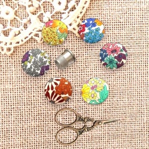 Pode incluir: Botões redondos cobertos de tecido com estampas florais, uma dedeira prateada e pequenas tesouras ornamentais. Os objetos estão dispostos sobre uma superfície de tecido bege texturizado, com renda no fundo.