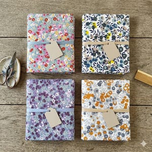 Peut inclure: Quatre paquets de tissu avec des motifs floraux aux couleurs variées, dont rose, bleu et orange. Chaque paquet est attaché avec un ruban ou une ficelle et porte une étiquette vierge. Des ciseaux et une petite coupelle sont visibles en arrière-plan.