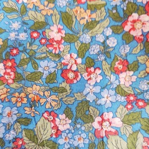 Könnte beinhalten: Nahaufnahme eines Stoffes mit floralem Muster. Der Stoff hat einen hellblauen Hintergrund mit einem sich wiederholenden Muster aus Blumen in Rot-, Rosa-, Orange- und Weißtönen mit grünen Blättern.